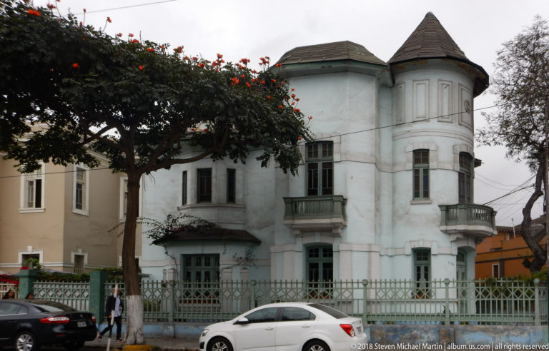 Pale blue historic corner residence with rounded turret, steep roof, balconies, tall windows, trees, and metal fence.