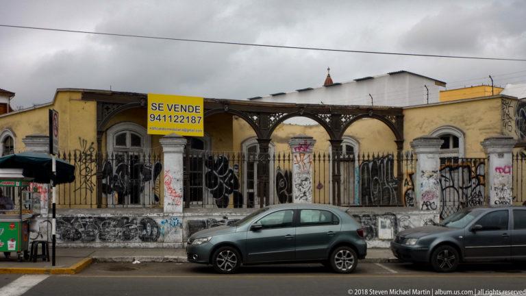 Weathered arched façade behind iron fencing with graffiti, metal canopy, parked cars, and yellow SE VENDE sign.