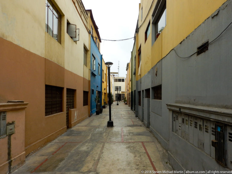 Narrow paved passage between ochre, blue, yellow, and gray buildings with overhead wires and a central streetlamp.