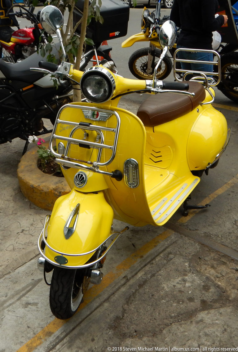 Bright yellow retro-style motor scooter with chrome racks, round headlamp, and brown seat parked at a curb.