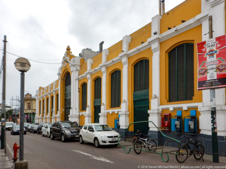Long ochre-and-white street façade with tall green louvered openings, arched entrance, parked cars, and bicycles.
