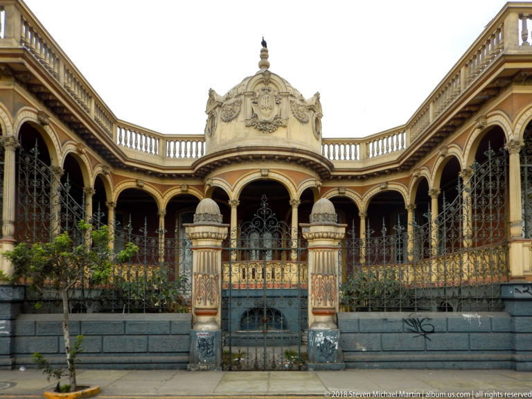 Centered ornate residence with sculpted crest, decorated entrance piers, arched bays, iron screens, and balustrade.
