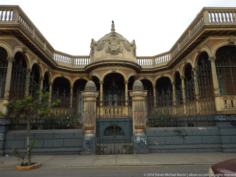 Ornate curving residence with repeated arches, iron screens, rooftop balustrade, central crest, and decorated gateposts.