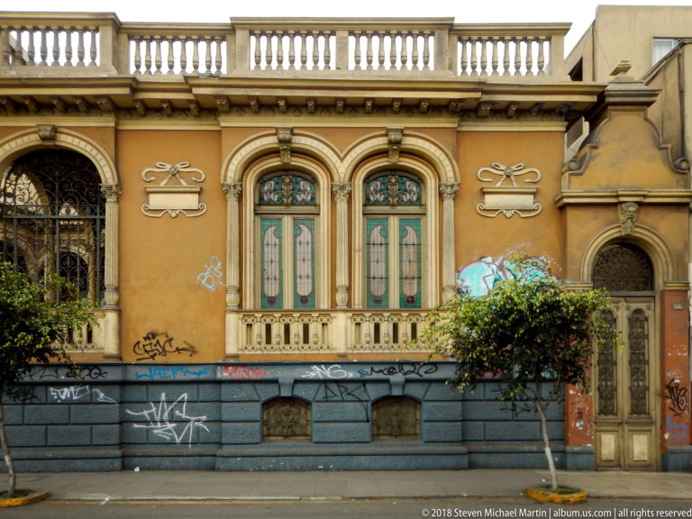 Ochre historic house with arched windows, decorative reliefs, roof balustrade, blue-gray base, and street graffiti.