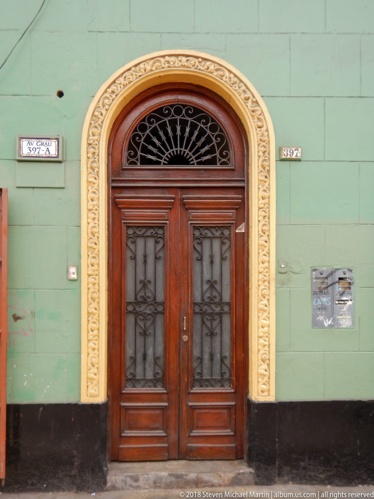 Dark wooden double door with ironwork and fan-shaped transom framed by cream ornament on a green wall.