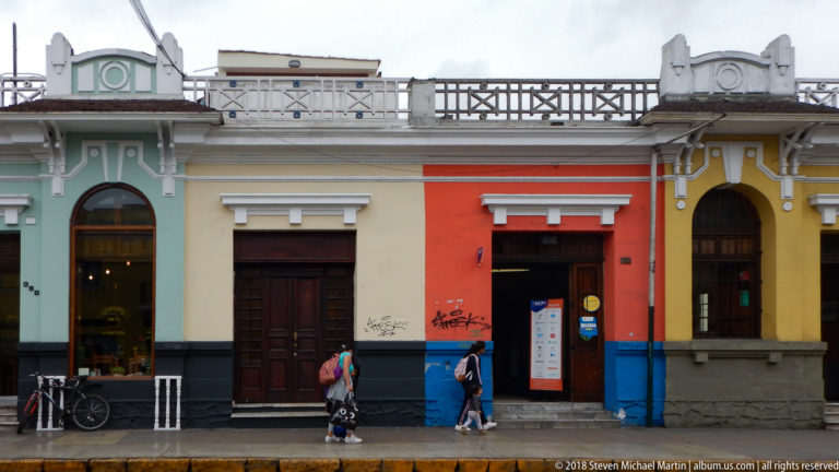 Mint, cream, orange, and yellow adjoining façades with dark bases, wooden doors, pedestrians, and a bicycle.