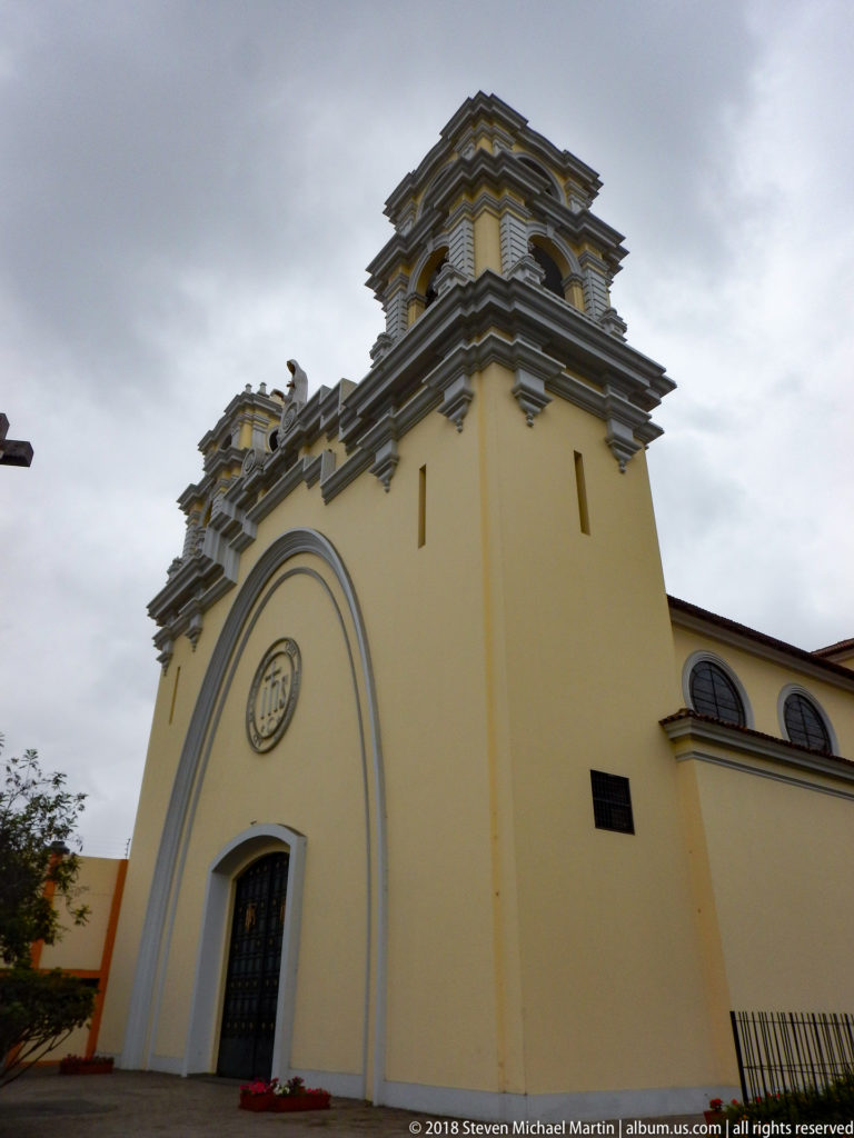 Low-angle view of yellow church with twin molded tower structures, arched façade, dark doorway, and circular IHS emblem.