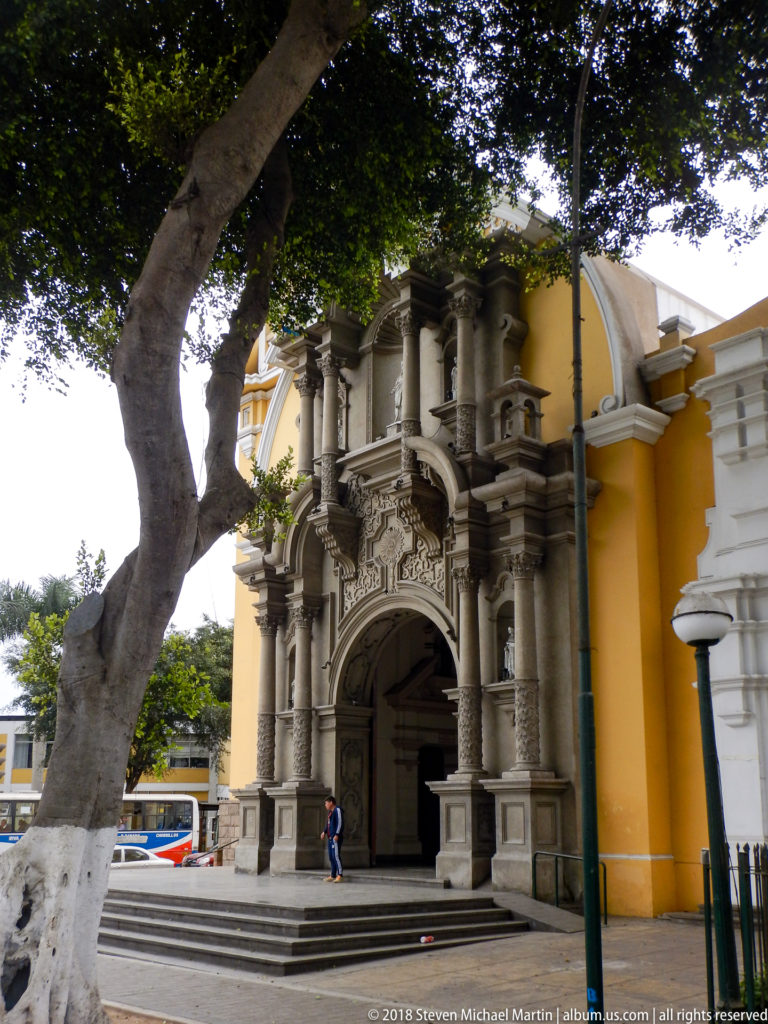 Oblique view of ornate gray church portal with columns, niches, broad steps, yellow wall, and large tree.