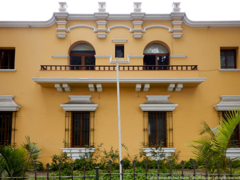 Yellow two-story façade with long balcony, arched upper windows, white trim, barred lower windows, and garden plants.