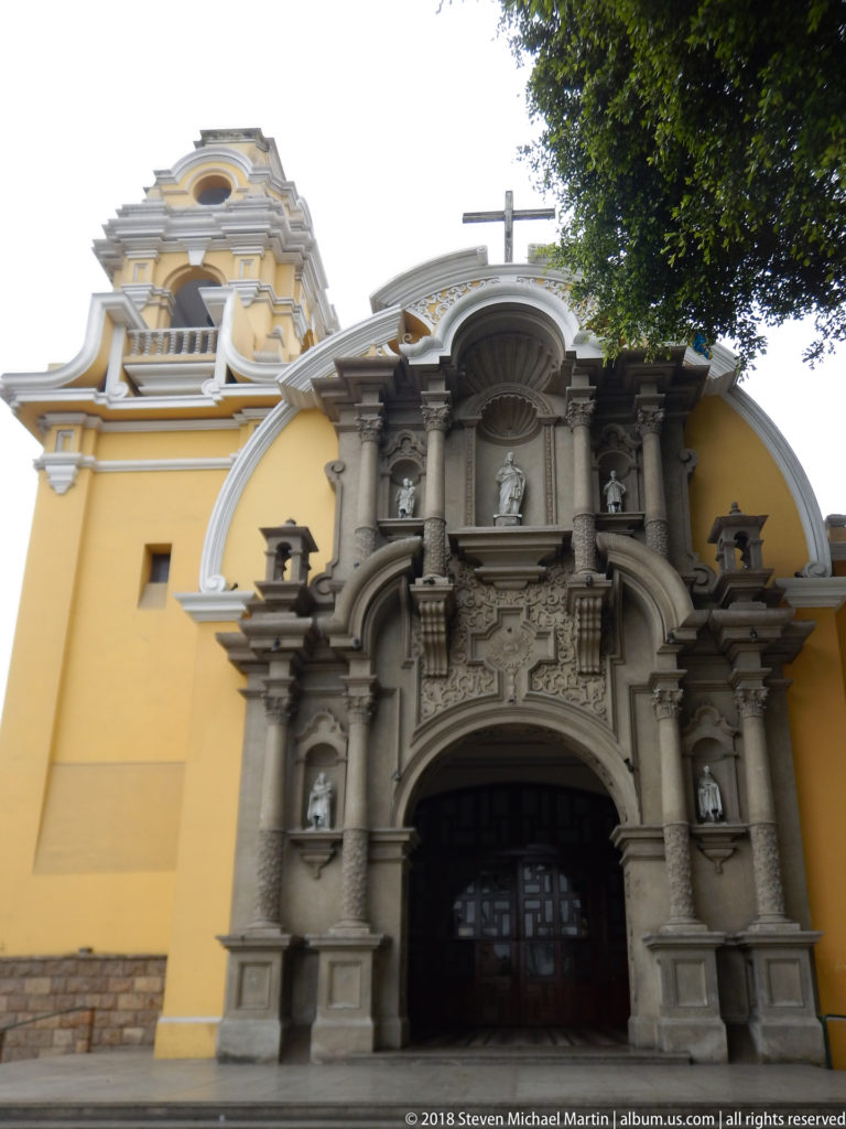 Ornate gray church portal with twisted columns, sculptural niches, arched doorway, and cross against yellow walls.