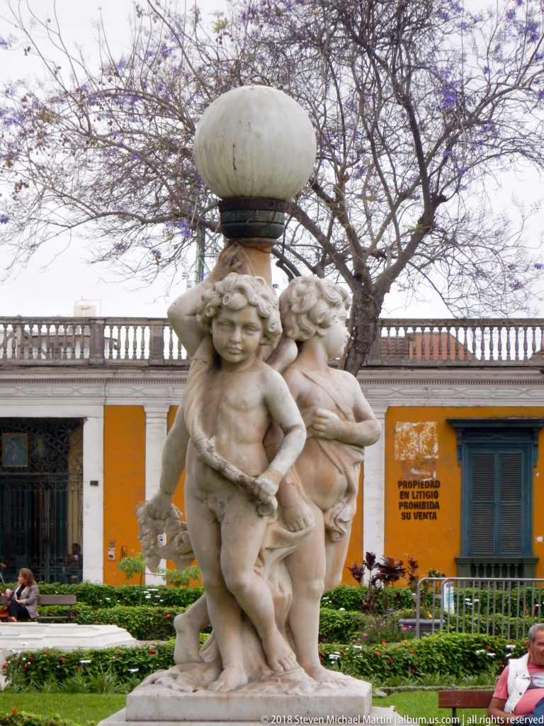 Stone-colored cherub figures clustered around a pedestal supporting a broad bowl in a landscaped public square.