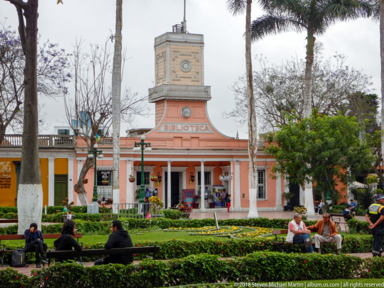 People seated in a landscaped park before the salmon-colored Biblioteca building and its square central tower.