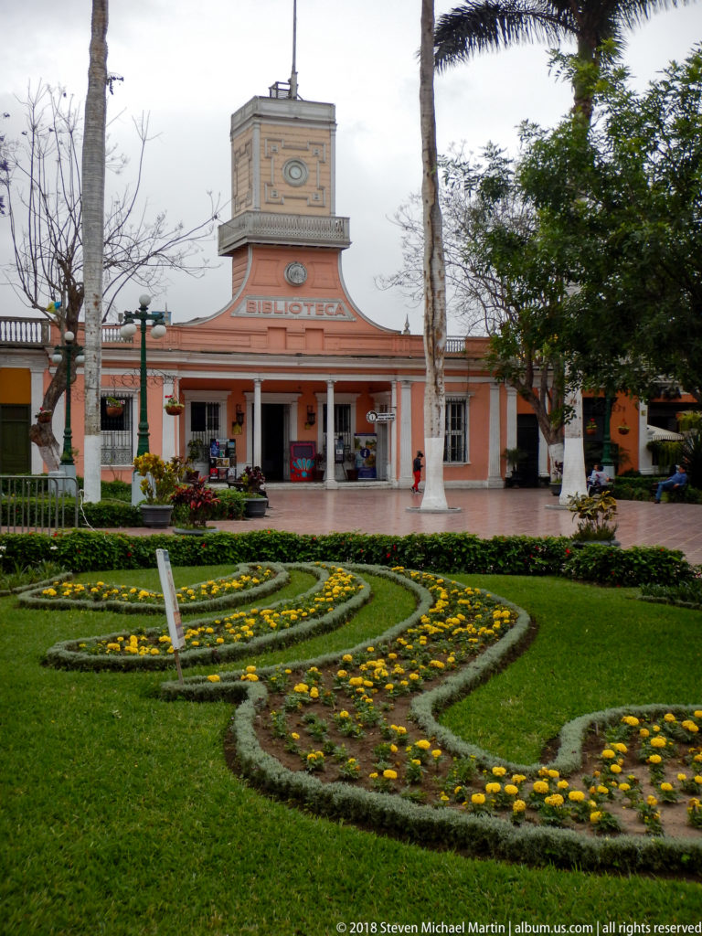 Formal lawn and yellow flower beds leading toward a salmon-colored Biblioteca building with central tower.