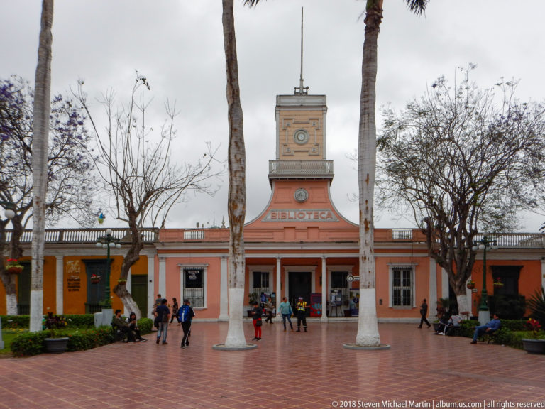 Frontal salmon-colored Biblioteca building with square central tower, tall palms, and people on a brick plaza.