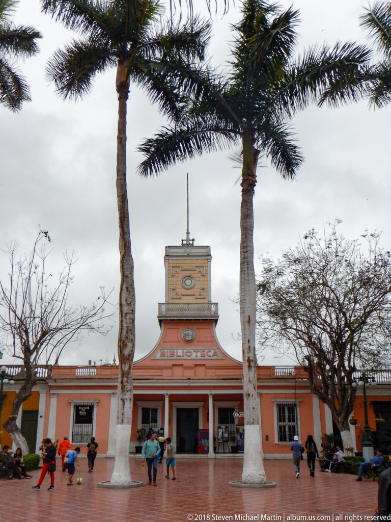 Salmon-colored building labeled BIBLIOTECA with central square tower, palms, and people crossing a brick plaza.