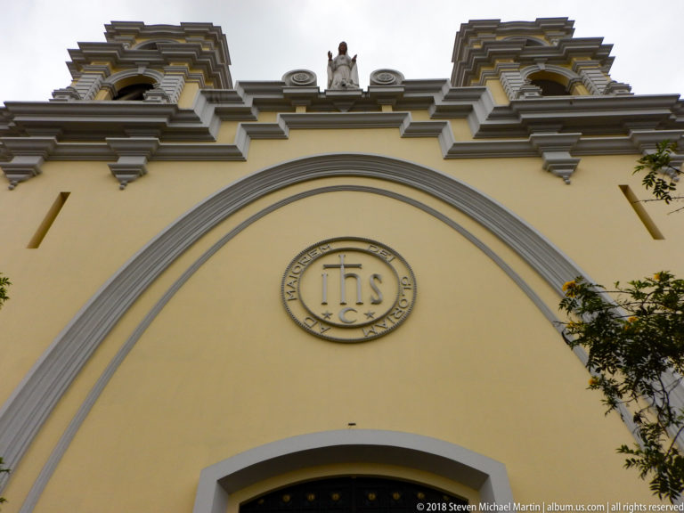 Yellow church façade with circular IHS Christogram, broad arched molding, layered cornices, and small roofline figure.