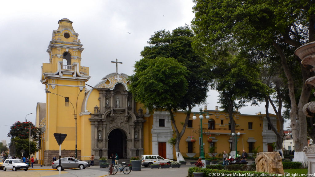 Yellow church with bell tower and ornate entrance beside mature trees, cyclists, pedestrians, and street traffic.
