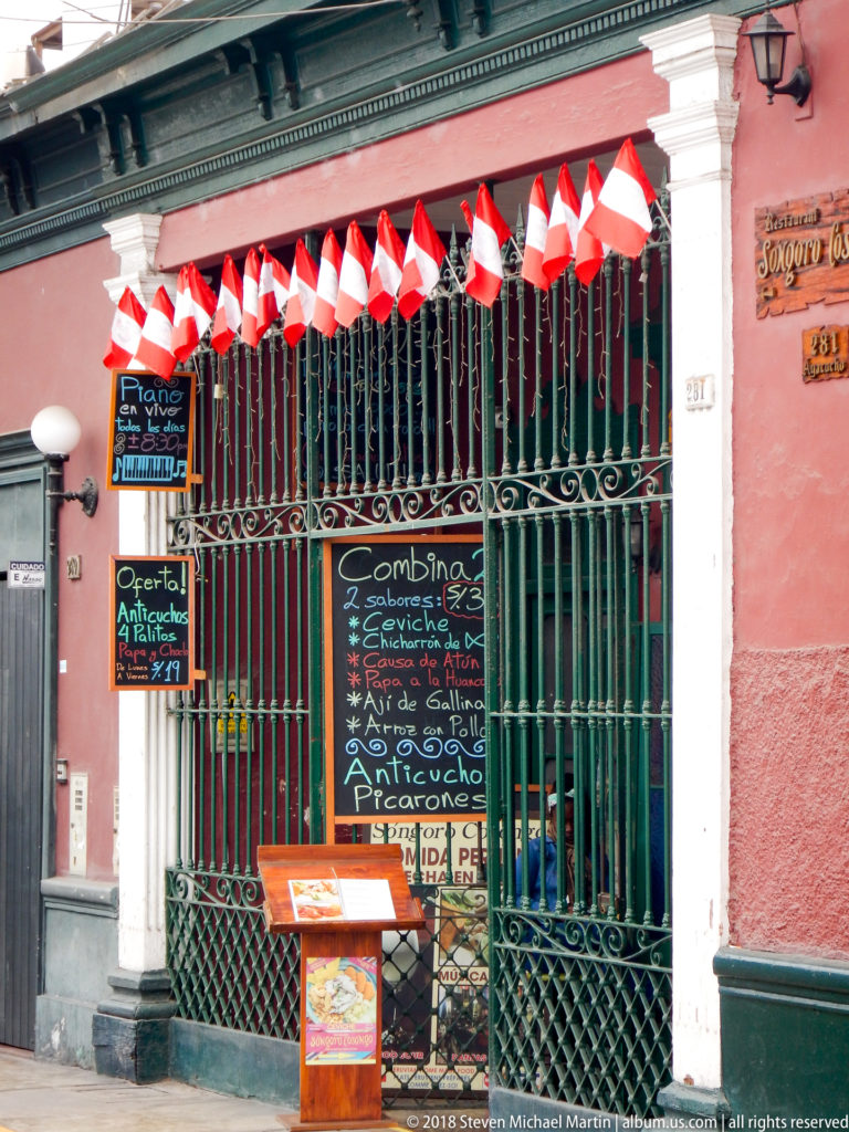 Restaurant entrance with green metal grilles, handwritten menu boards, and a row of red-and-white Peruvian flags.