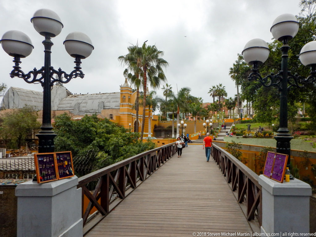 Wooden Puente de los Suspiros with cross-braced rails, globe lamps, pedestrians, palms, and yellow buildings ahead.