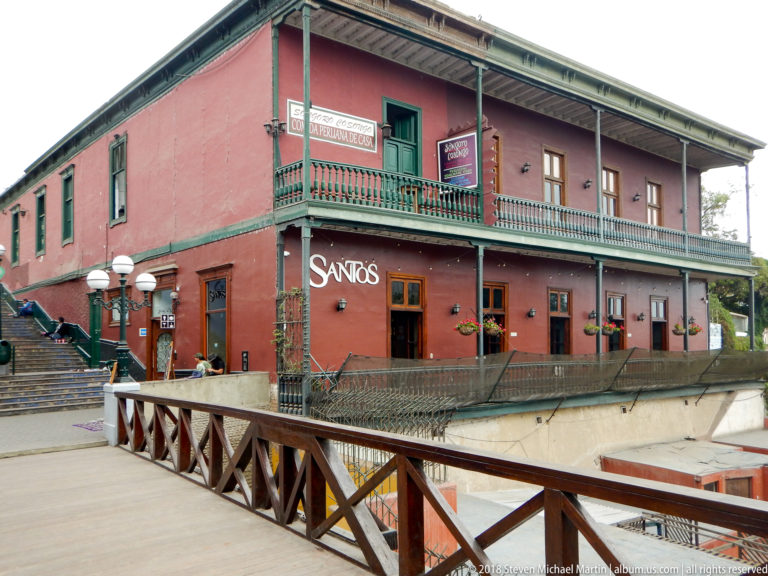 Red two-story building with green balcony rails, deep eaves, signage, and flower boxes beside a wooden bridge.