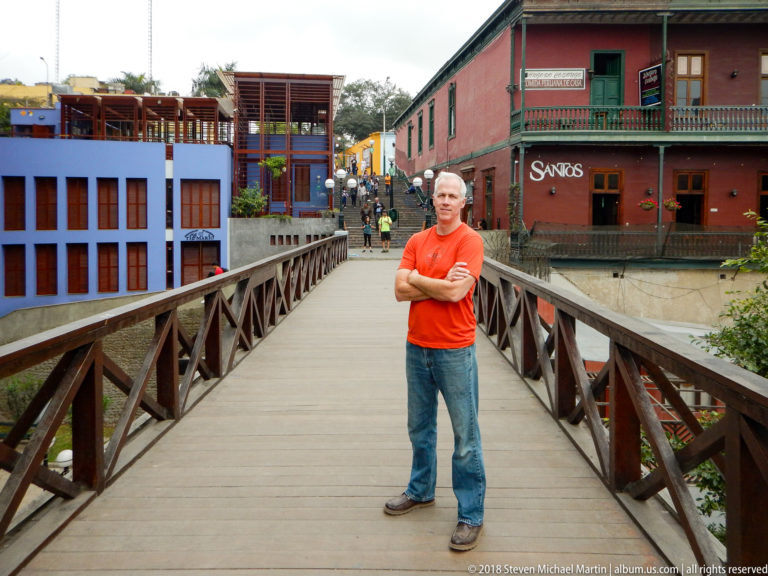 Man in orange shirt standing on the wooden Puente de los Suspiros with colorful buildings and pedestrians beyond.
