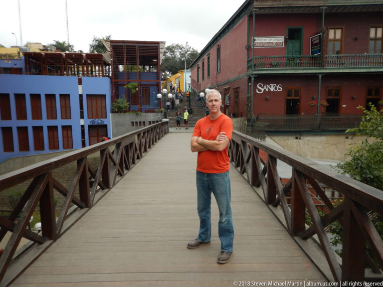 Full-length man in orange shirt centered on a wooden pedestrian bridge with colorful neighborhood buildings behind.