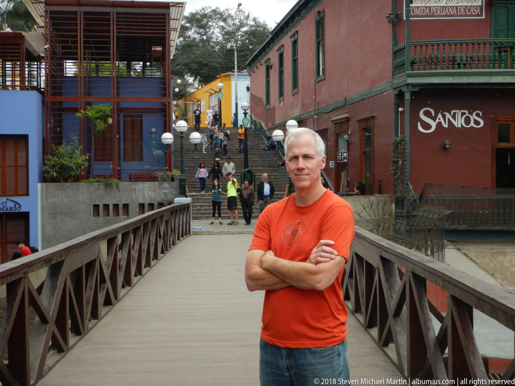 Man in an orange shirt standing with folded arms on a wooden pedestrian bridge amid colorful Barranco buildings.