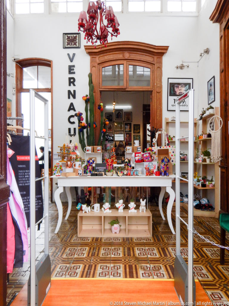 Bright craft shop interior with colorful small objects on a central table, wall shelves, wood trim, and patterned tile floor.