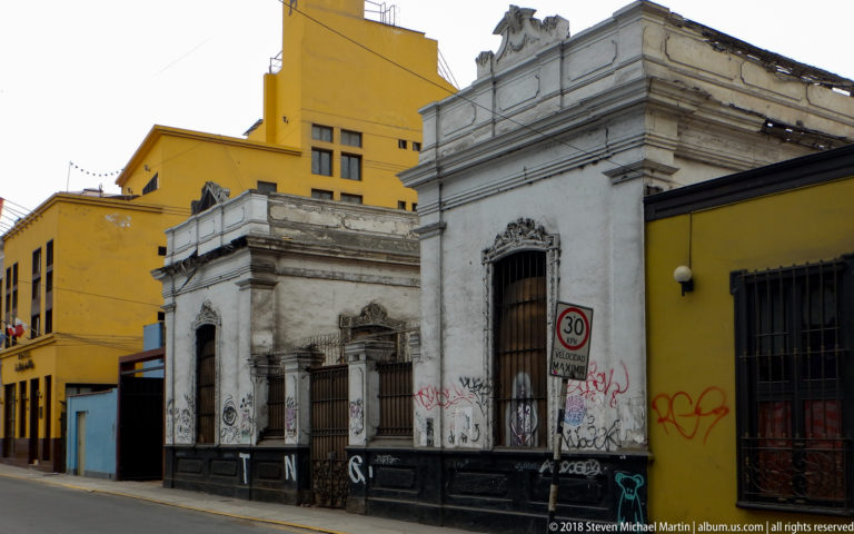 Weathered ornate white historic façades with graffiti beside a taller yellow building on a Barranco street.