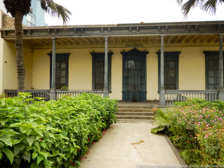 Pale yellow historic building with long columned veranda, central steps, dark doors, and lush garden beds.