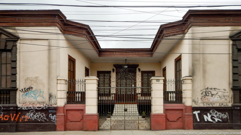Symmetrical cream-and-red historic façade with recessed iron-gated entrance, dark wood doors, graffiti, and overhead wires.