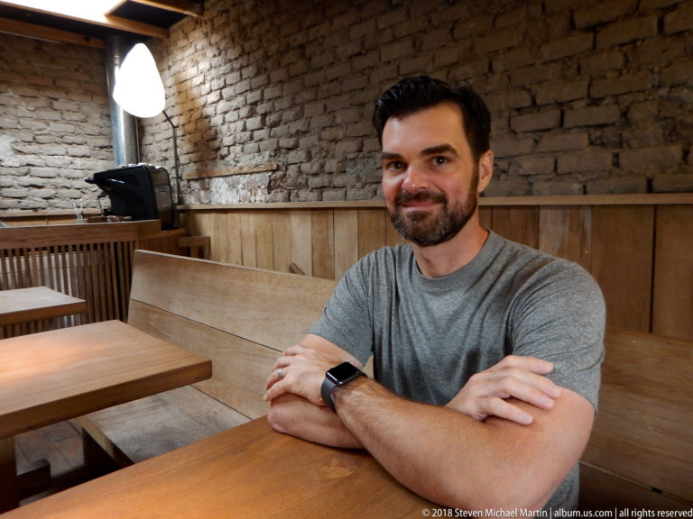 Bearded man in a gray shirt seated with folded arms at a wooden table against an exposed brick wall.