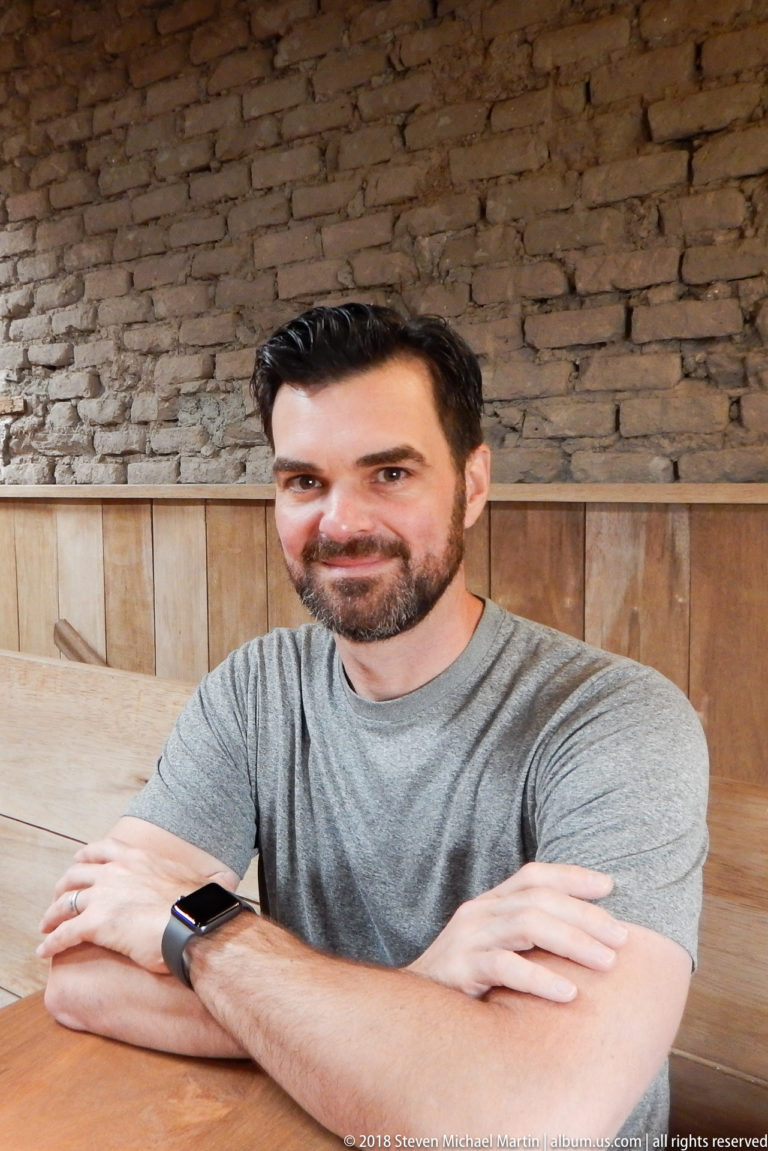 Man in a gray shirt seated with crossed arms at a wooden restaurant table against brick and wood walls.