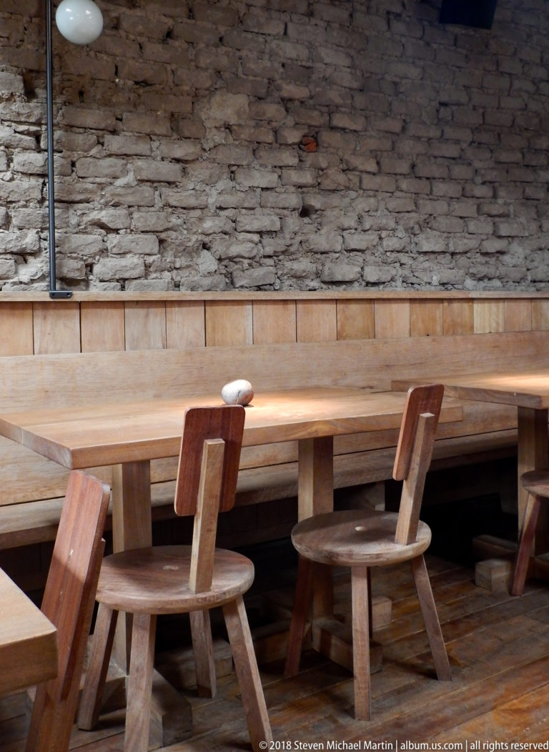 Empty rustic restaurant seating with heavy wooden tables and chairs against exposed brick and metal ductwork.