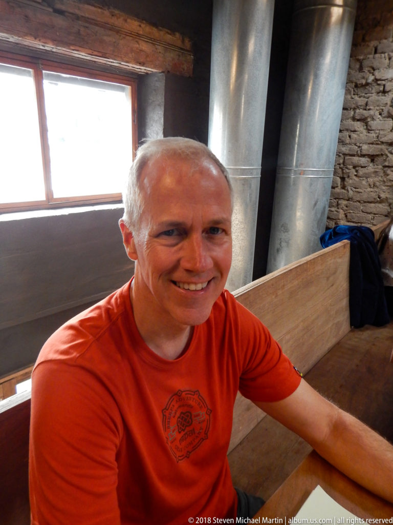 Smiling man in an orange shirt seated at a wooden table with brick walls and metal ducts behind him.