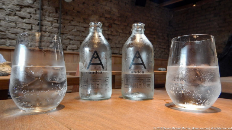 Sparkling-water glasses in the foreground framing two chilled andea bottles against a brick restaurant wall.