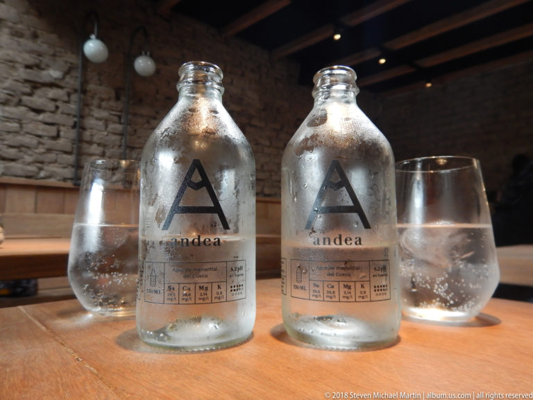 Two chilled glass bottles labeled andea with water-filled tumblers on a wooden restaurant table.