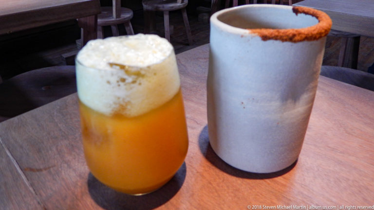 Orange foamy drink beside a tall gray ceramic cup with a reddish seasoned rim on a wooden table.