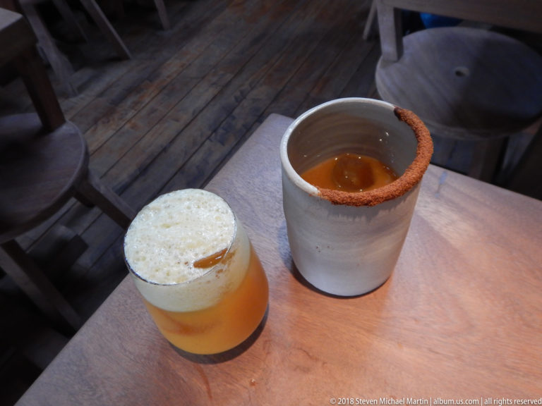 Two restaurant drinks, one amber with a foamy top and one in a ceramic cup with a reddish spiced rim.