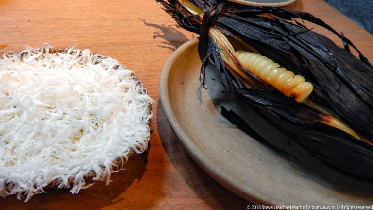 Large pale corn kernels in a blackened husk beside a mound of finely shredded white garnish.
