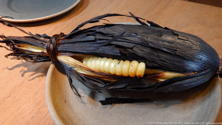 Large pale corn kernels exposed inside a heavily blackened roasted husk on a plate.