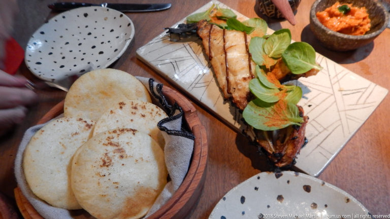 Restaurant table with round flatbreads, a long grilled dish topped with greens, and a small bowl of reddish condiment.