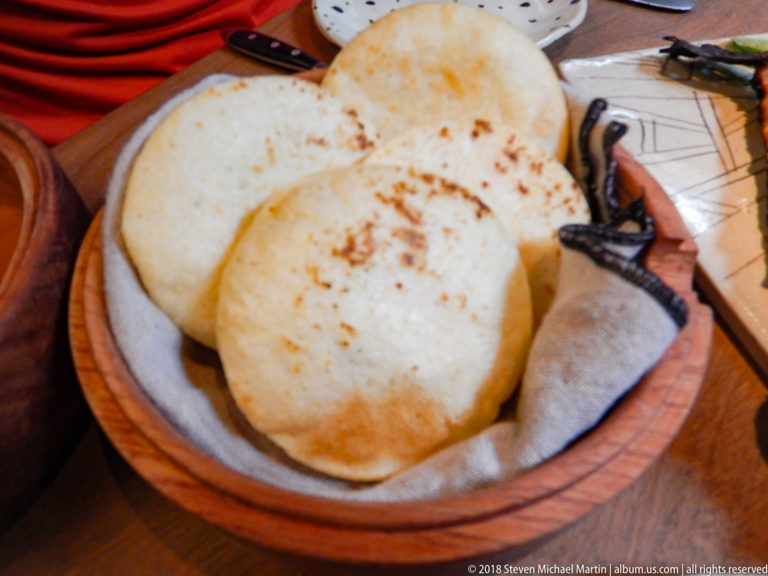 Four lightly browned round flatbreads stacked in a cloth-lined wooden serving bowl on a restaurant table.