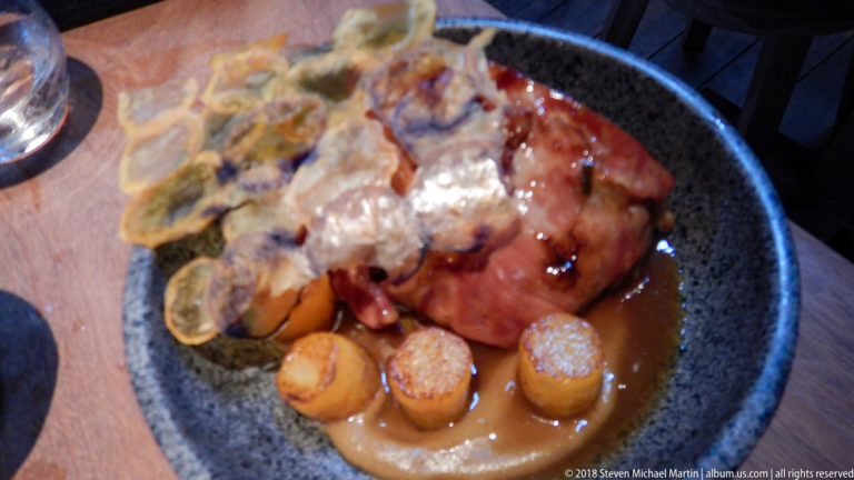 Glazed roasted meat in a dark bowl with brown sauce, small cylindrical vegetables, and crisp garnishes.