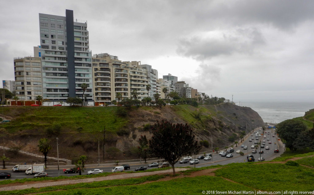 High-rise apartments above a steep green cliff and busy curving coastal highway under an overcast sky.