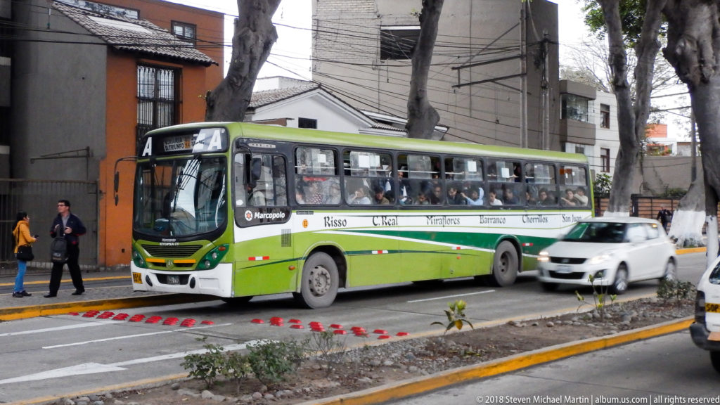 Green-and-white city bus traveling past pedestrians, trees, utility lines, and low-rise buildings on a Barranco street.
