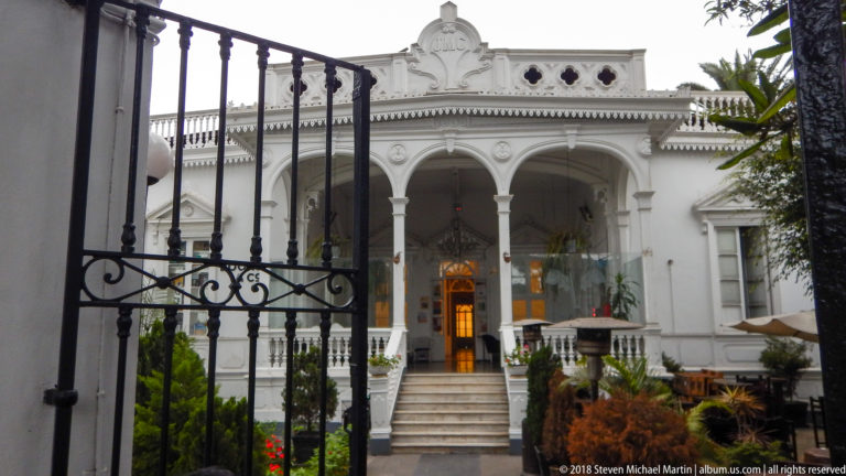 White historic house with raised arched veranda and central steps, viewed through a black wrought-iron gate.