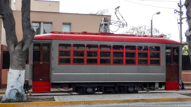 Preserved gray-and-red electric streetcar with many windows and roof-mounted electrical apparatus displayed beside the road.