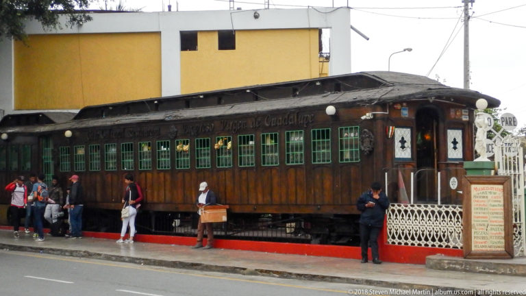 Dark wooden railcar with green-trimmed windows beside a street platform, with visitors and restaurant signage.