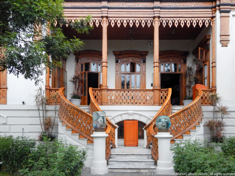 Raised wooden veranda with carved posts, ornate railings, arched windows, and symmetrical curved staircases.